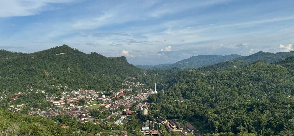 Panoramic view of Sawahlunto, a historic town in West Sumatra, Indonesia, surrounded by lush green hills and colonial-era buildings under a blue sky.