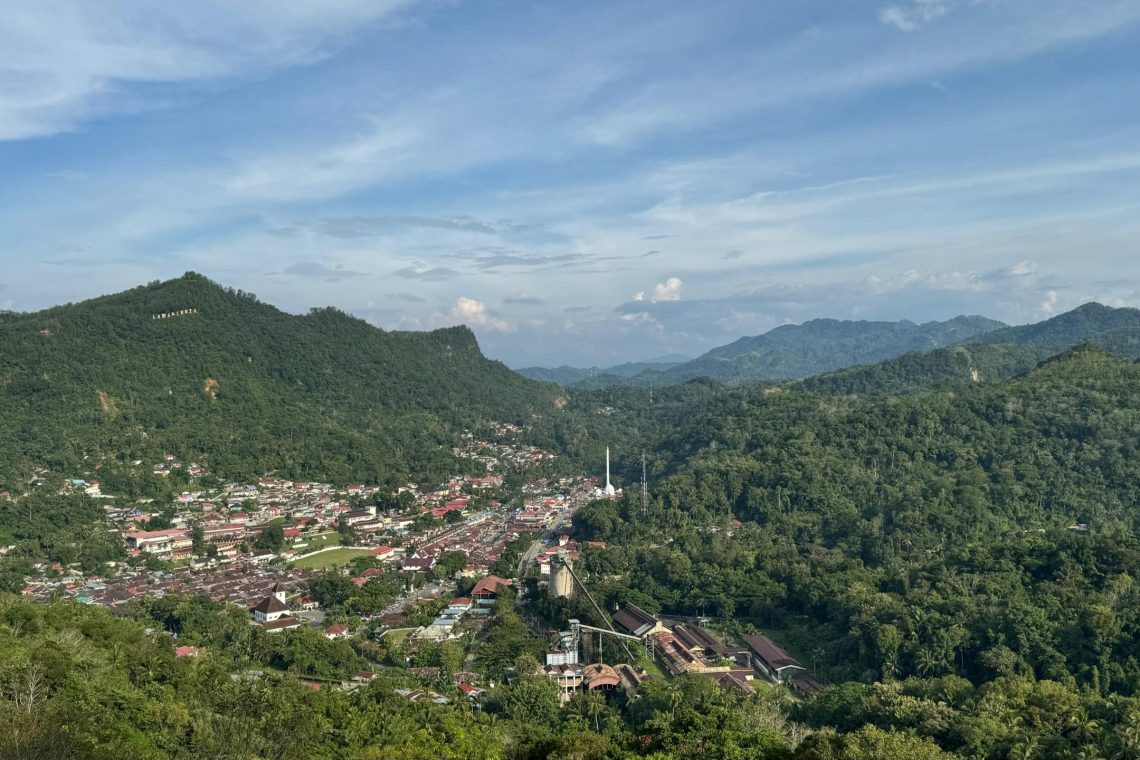 Panoramic view of Sawahlunto, a historic town in West Sumatra, Indonesia, surrounded by lush green hills and colonial-era buildings under a blue sky.